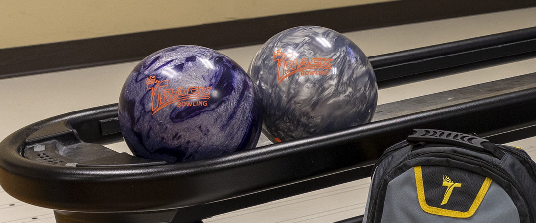 Two Track Bowling balls on ball rack with the top of a backpack seen next to them.